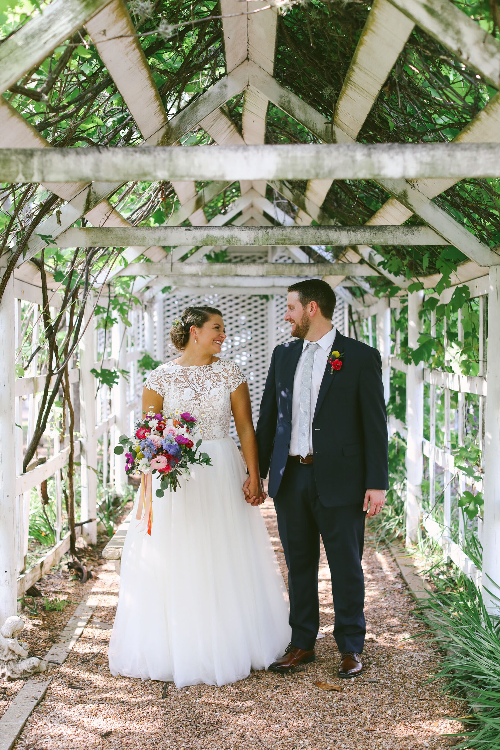 A bride and groom stand under a wooden arbor covered in greenery, holding hands and smiling at each other. The bride wears a white lace dress and holds a colorful bouquet; the groom wears a dark suit with a floral boutonniere.