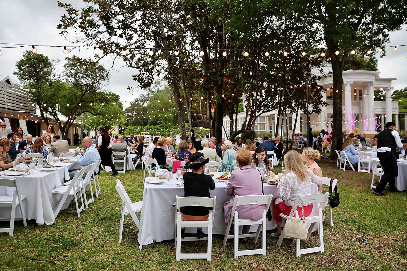 Guests sit at round tables with white tablecloths, dining outdoors under string lights and trees, near a large white building. The atmosphere is festive and elegant.