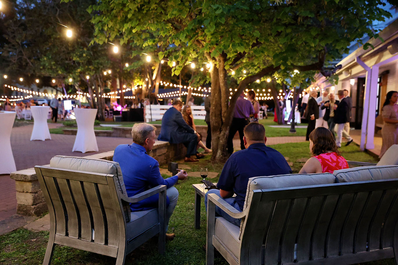 People sit on outdoor benches at a nighttime garden event, with string lights hanging above, cocktail tables scattered around, and guests mingling in the background under trees and soft lighting.