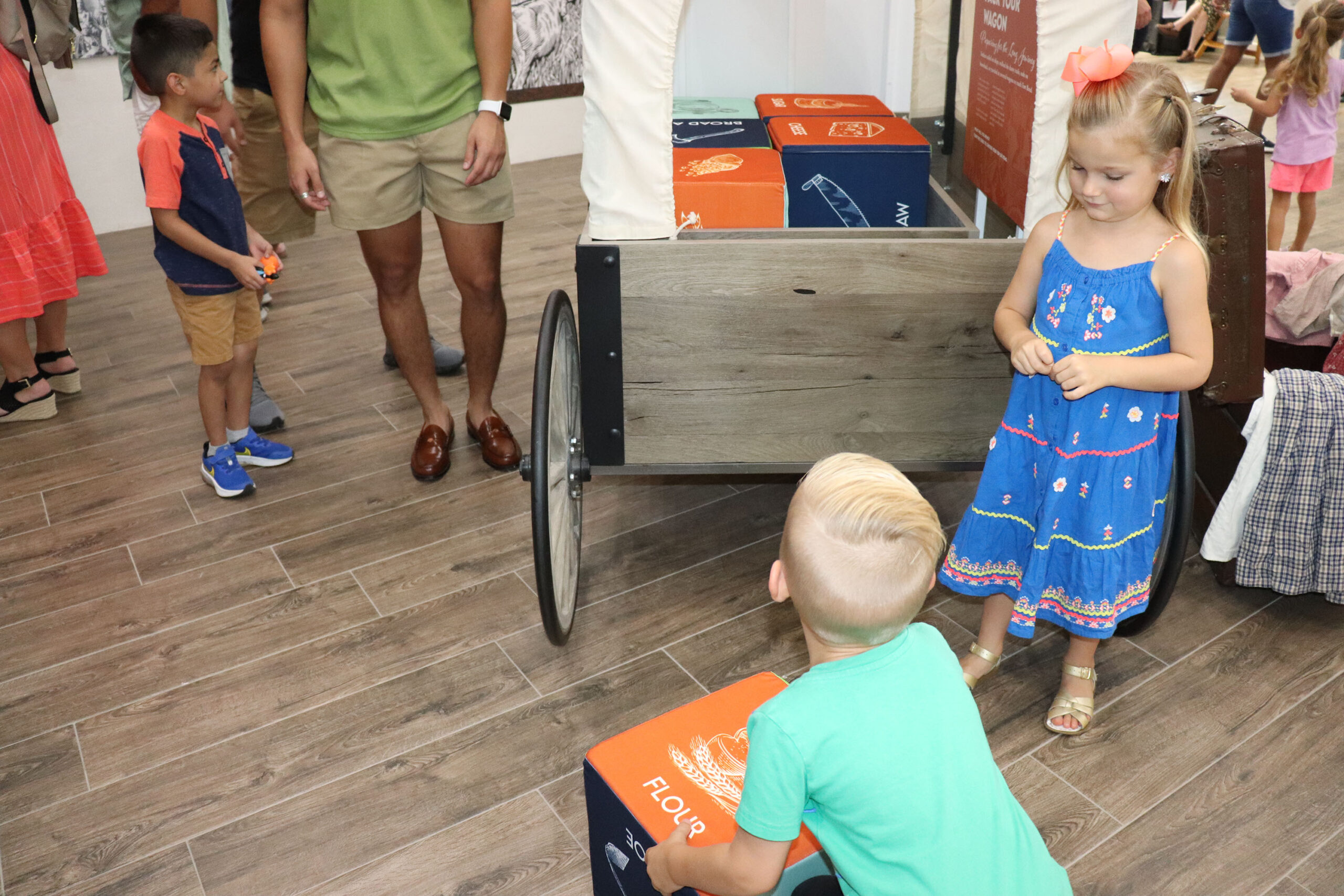 Several young children stand near a wooden wagon filled with colorful boxes indoors. A girl in a blue dress faces a boy in a green shirt, with adults and other children nearby on a wood floor.