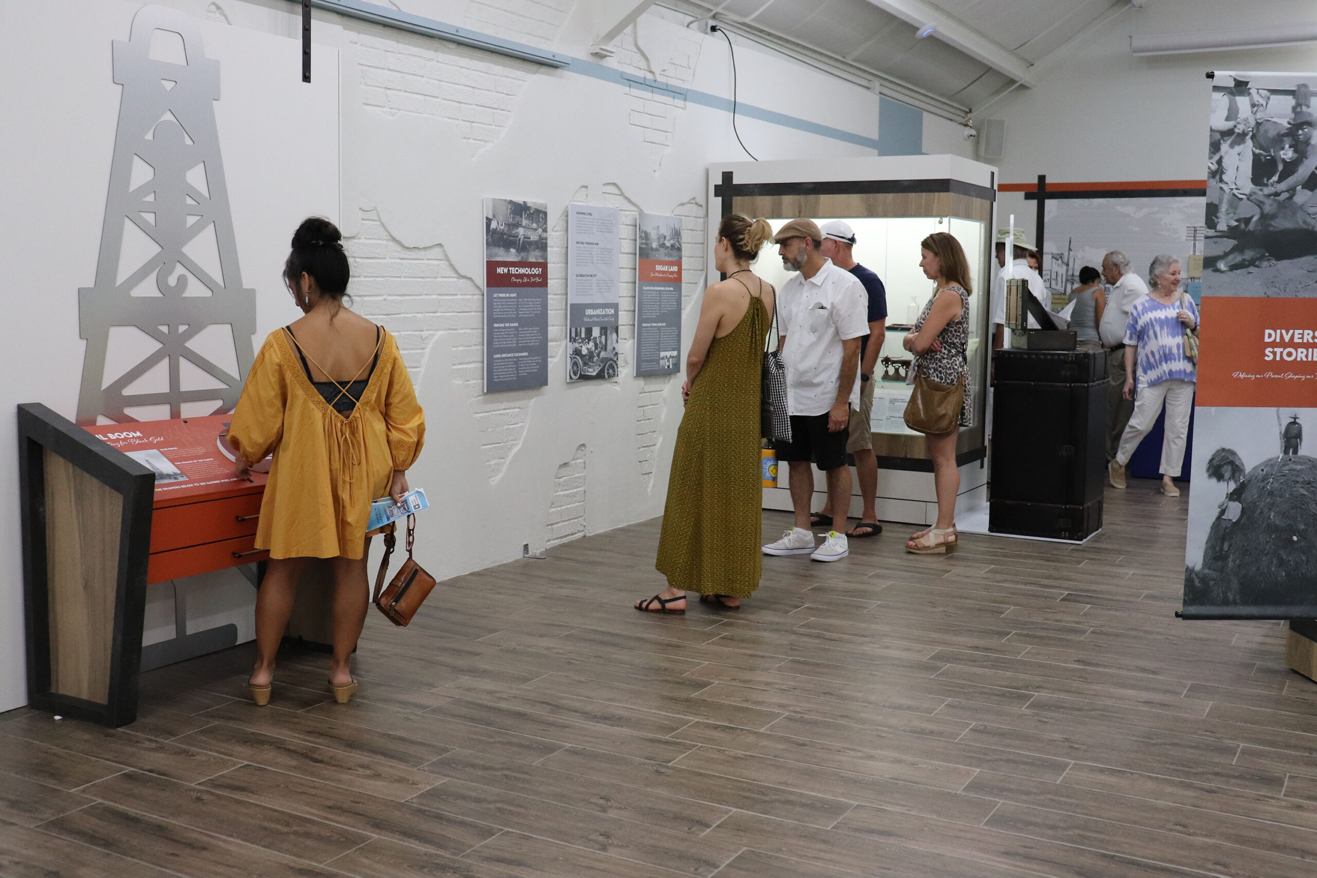 People explore a museum exhibit with informational displays about oil, including a large oil derrick graphic. One woman interacts with a touchscreen while others read wall-mounted panels. The room has wood flooring and white brick walls.