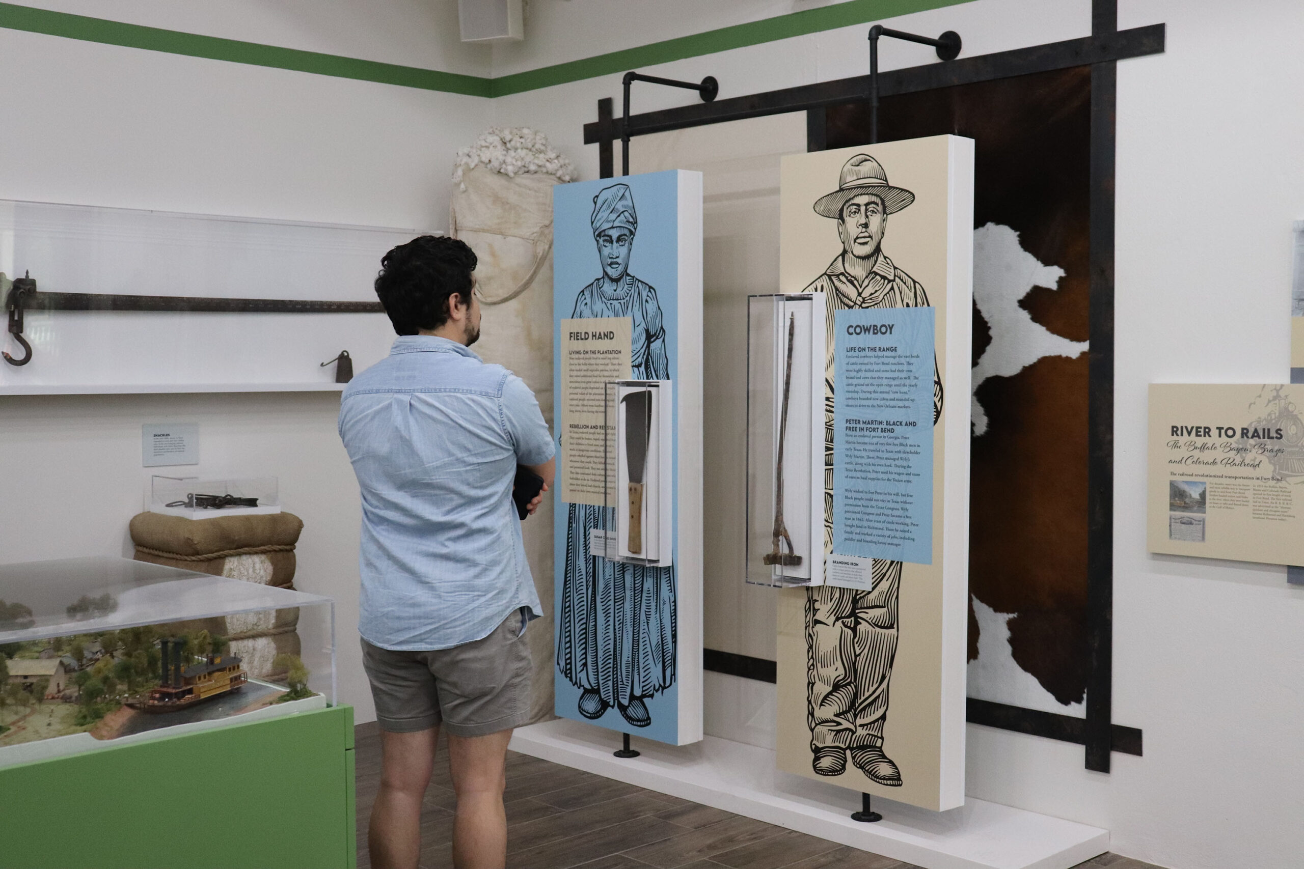 A person in a light blue shirt and shorts stands in a museum, facing two tall displays featuring illustrations and information about a freed hand and a cowboy, with historical artifacts between them.