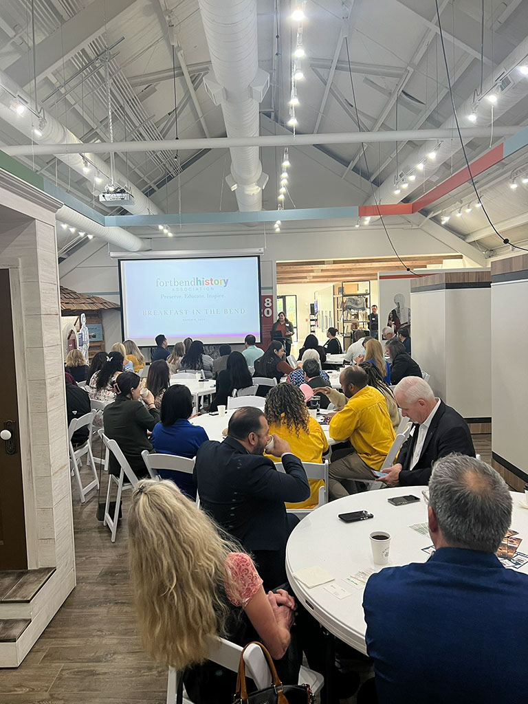 A group of people sit at round tables, facing a large screen displaying the words "fortbendhistory breakfast & business," inside a bright, modern room with high ceilings and exposed ducts.