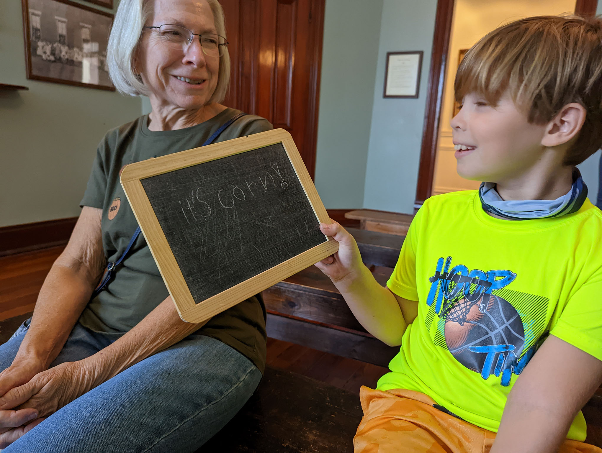 A smiling boy in a neon yellow shirt holds a small chalkboard with “It’s candy” written on it, sitting next to an older woman with glasses in a room with wooden floors and walls.