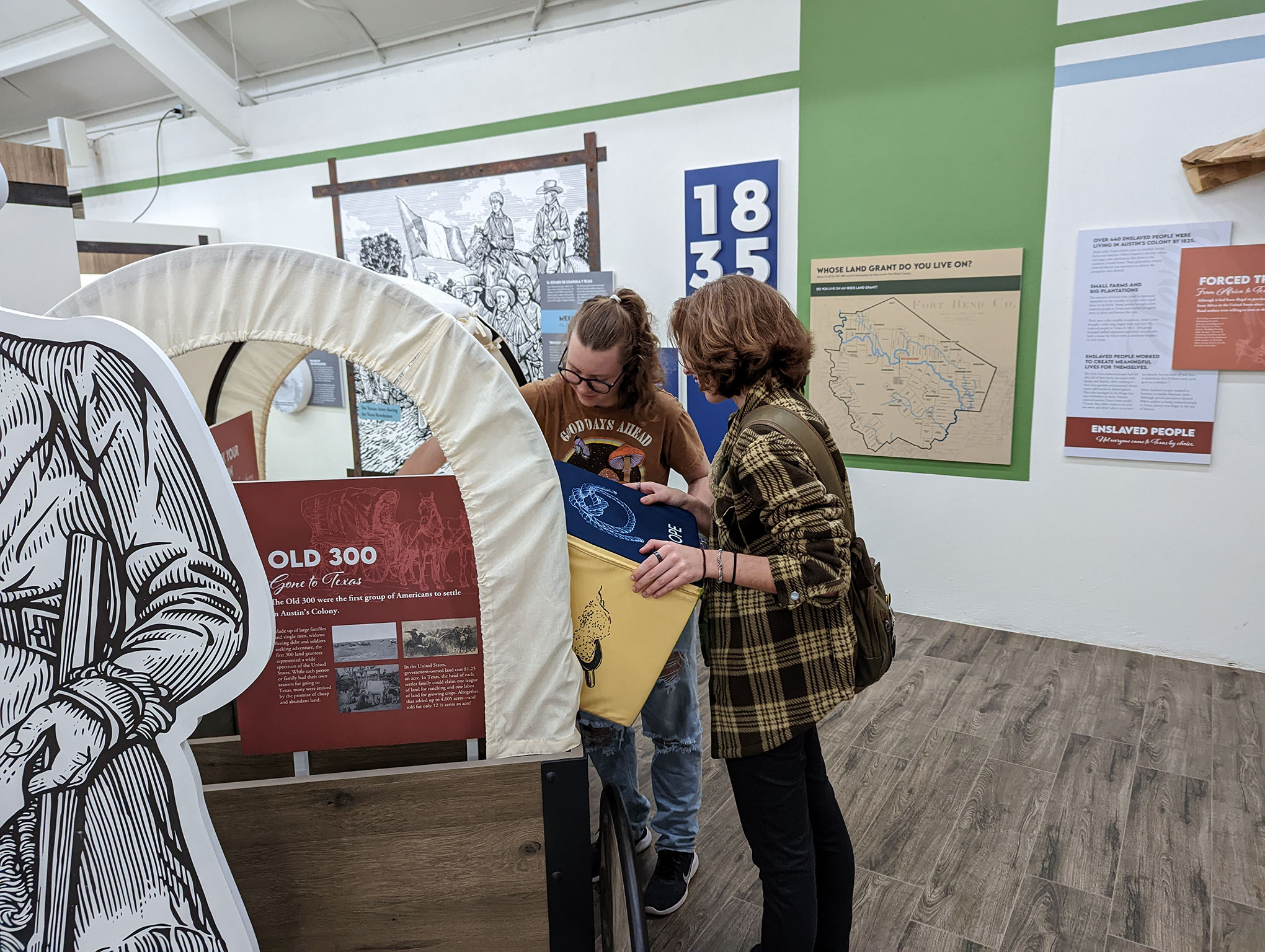 Two people examine artifacts in a covered wagon exhibit at a museum. They are looking closely at items inside, with educational displays, maps, and historical timelines visible on the surrounding walls.