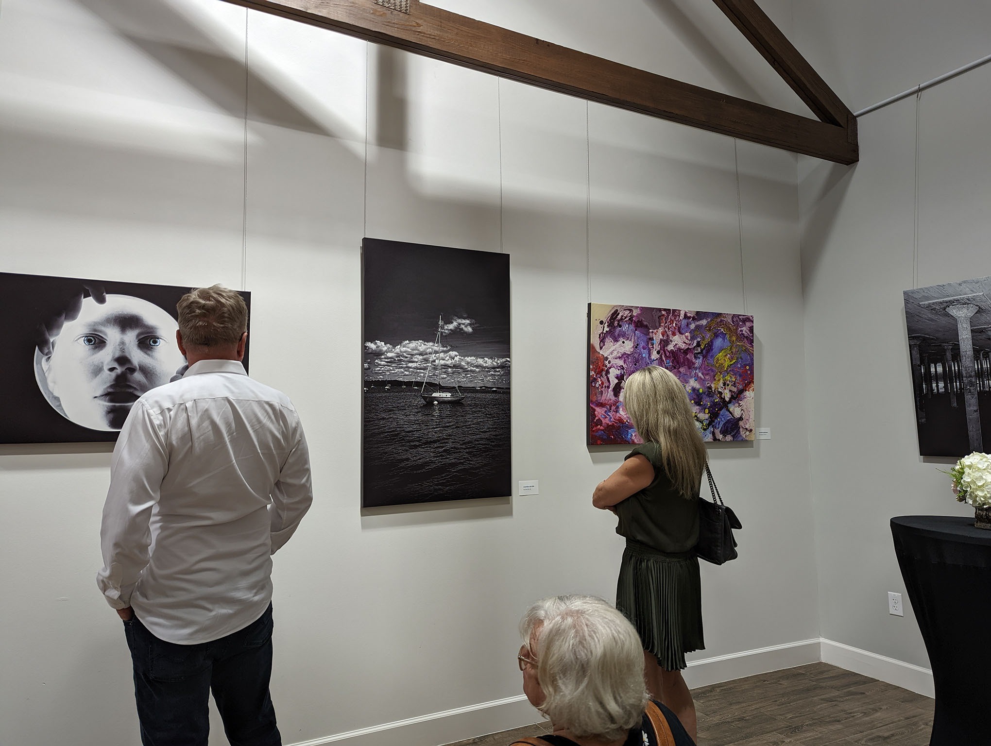 Three people view artwork in a modern gallery. Two stand observing black-and-white and colorful abstract paintings on the wall; one person is seated in the foreground, facing away. The room has white walls and wooden beams.