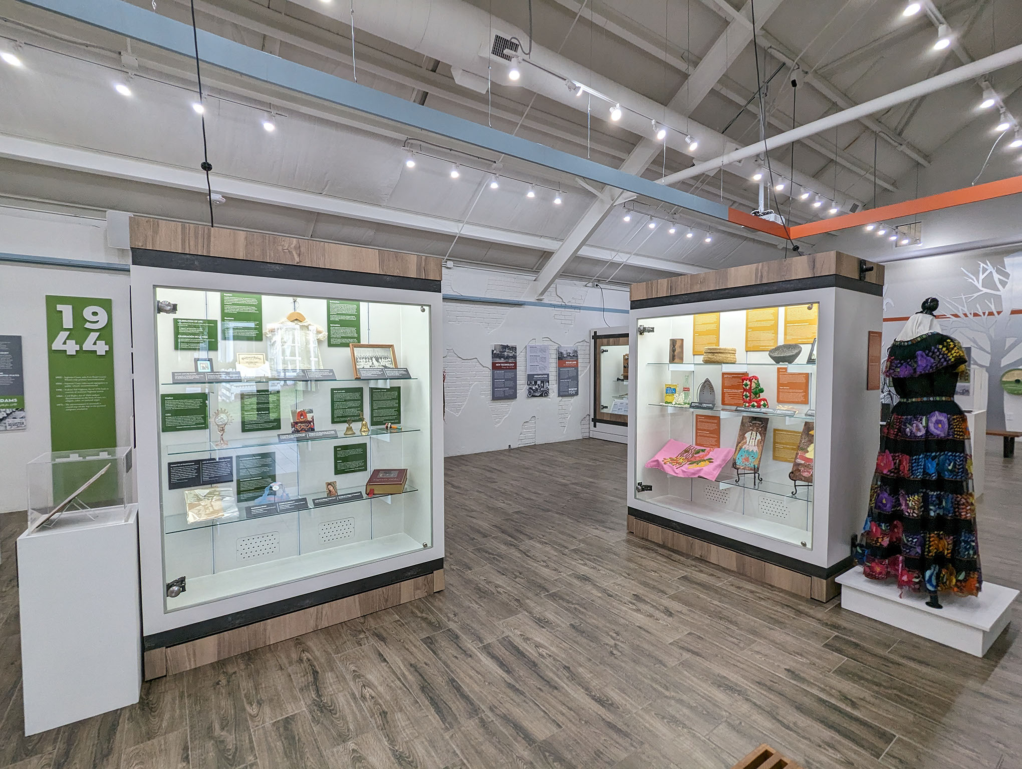 A museum exhibit features two large glass display cases with books, artifacts, and documents. To the right, a colorful dress made of fabric squares is displayed on a mannequin. The room has white walls and a wood floor.