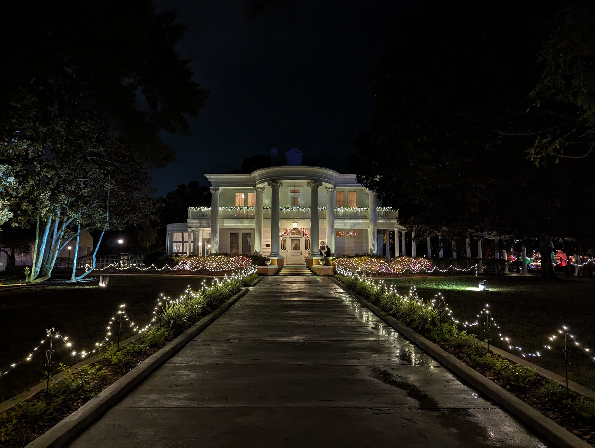 A grand white mansion with columns is illuminated at night, decorated with string lights along the walkway and bushes, creating a festive and welcoming atmosphere. Trees frame the house on both sides.