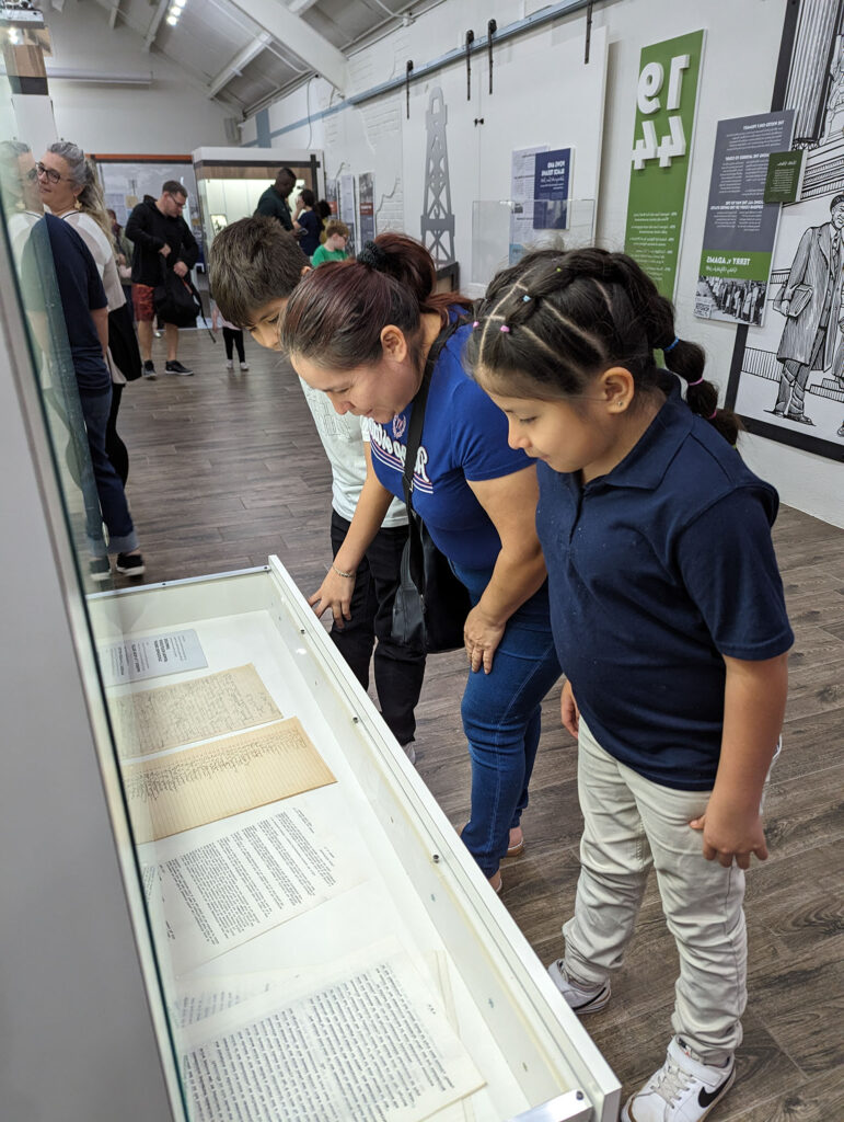 A woman and two children look at historical documents displayed in a glass case at a museum, with several other visitors in the background.