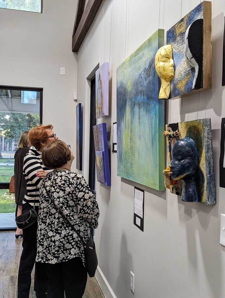 Two women closely view mixed-media and sculptural artwork displayed on a gallery wall, with natural light streaming in from large windows on the left.