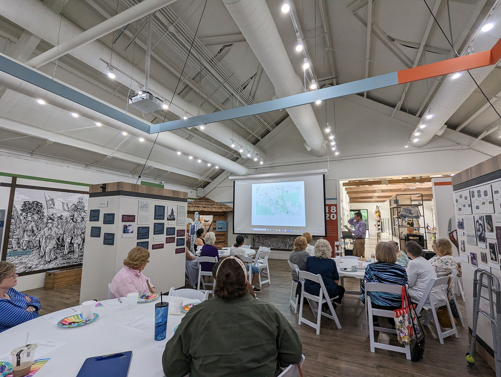 A group of people sit at round tables in a well-lit room, watching a presenter in front of a large projection screen displaying a map. Display boards and historical exhibits line the walls.