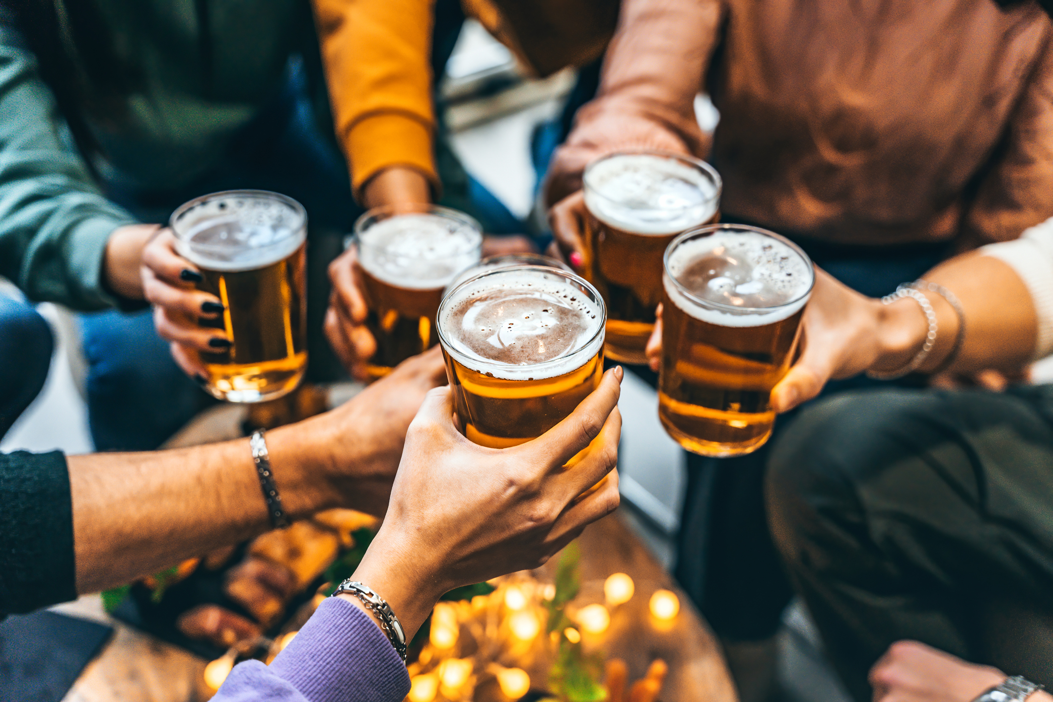 A group of people clink glasses of beer together in a toast, seen from above. The table below has some candles and food, and everyone is wearing casual, colorful clothing.