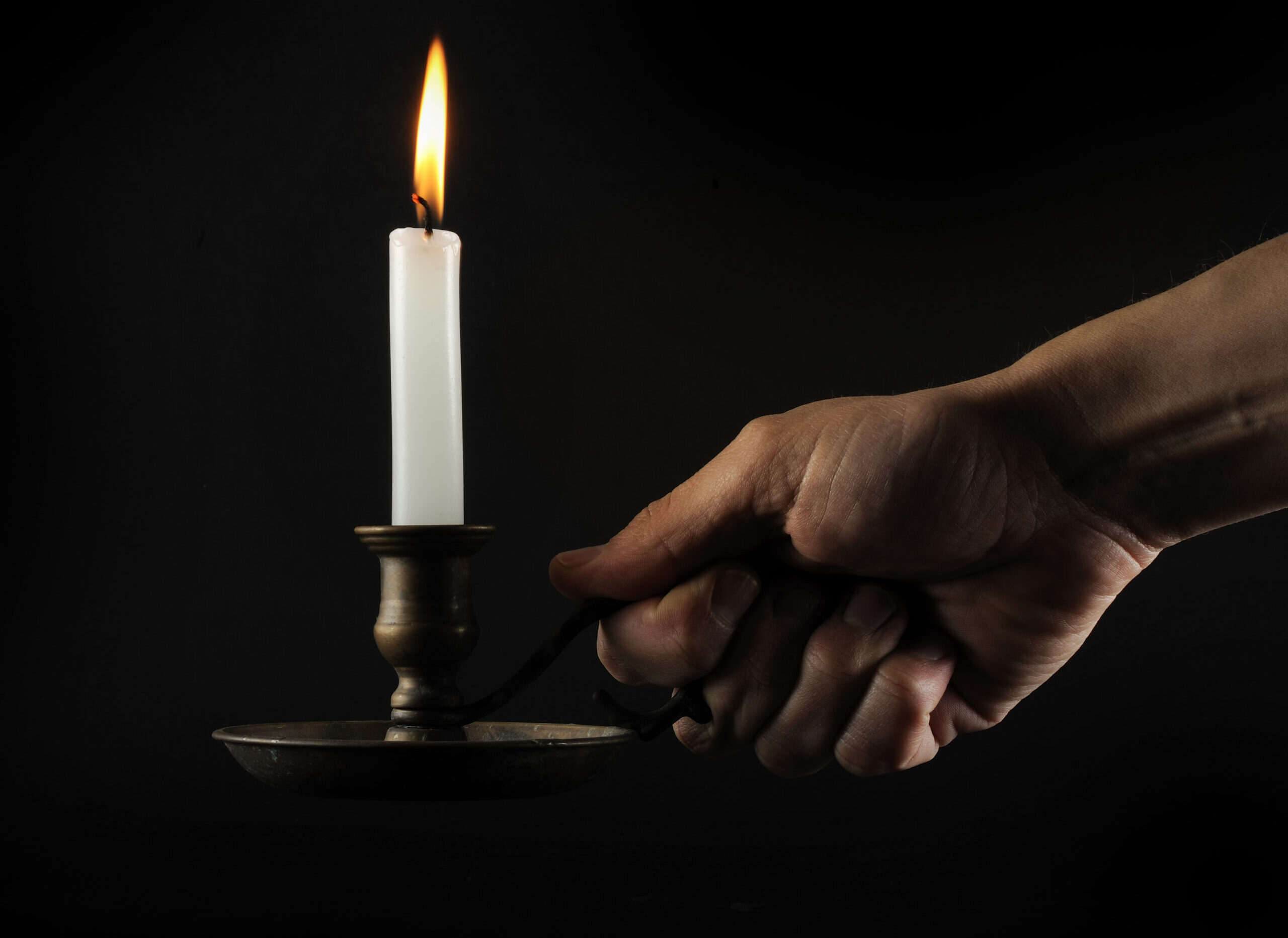 A hand holds a brass candle holder with a lit white candle against a black background, the flame casting a warm glow.