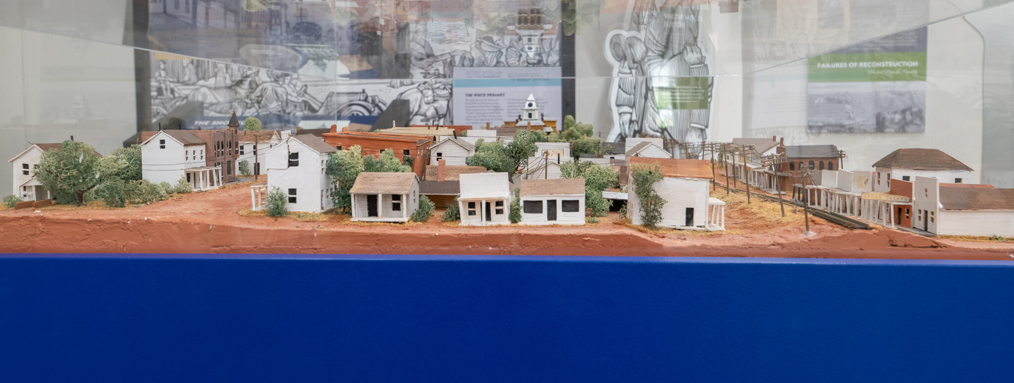 A detailed miniature diorama of a small town with white wooden buildings, dirt roads, and trees, displayed behind glass with informational posters in the background.