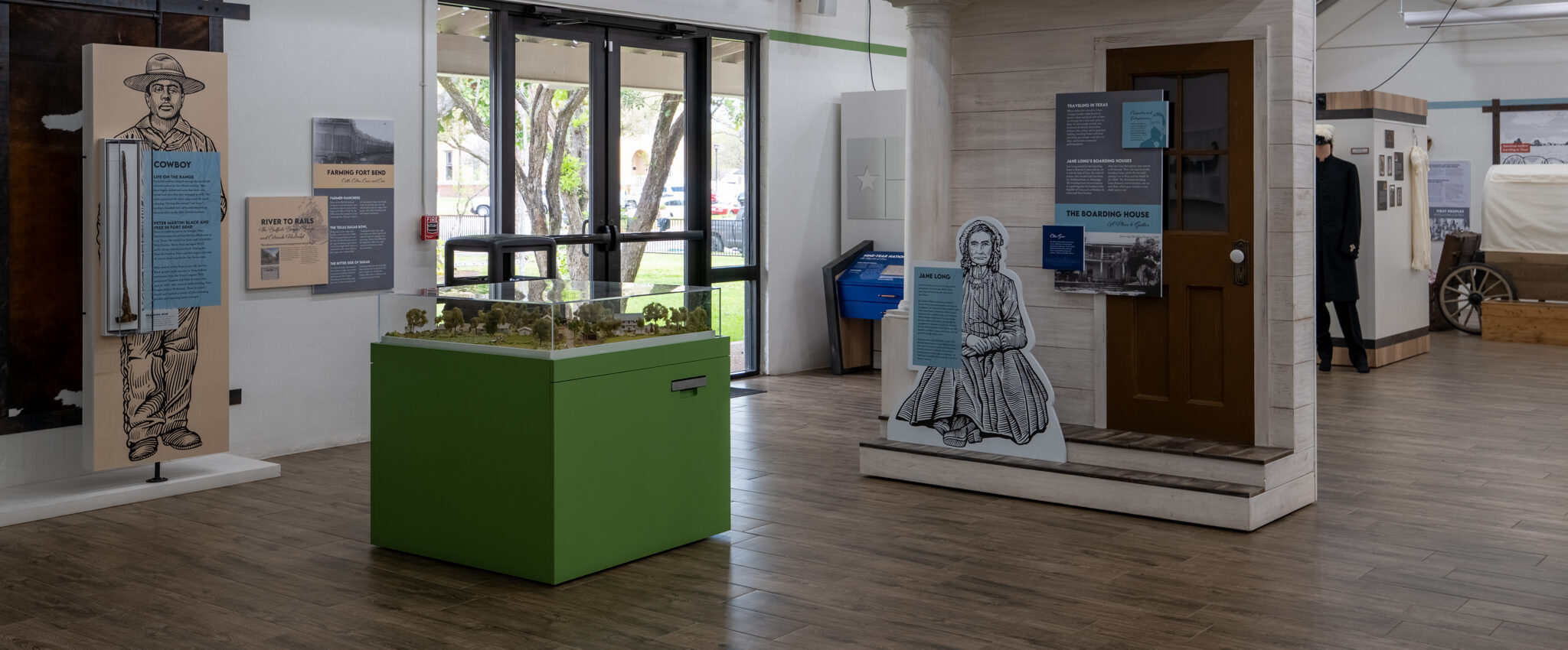 A museum exhibit features life-size cutouts of historical figures, informational panels, a display case on a green pedestal, and various artifacts in a well-lit room with large windows.