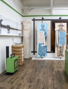 Museum exhibit with illustrated panels of a field hand and a cowboy, cotton bales, a large wrapped bundle, a green box, and an old farming tool displayed on the wall in a glass case.