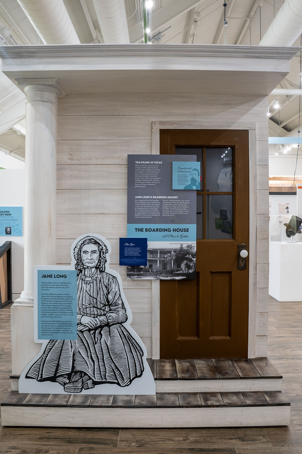 A museum exhibit features a small wooden house facade with two columns, a brown door, and informational signs about Jane Long. A black-and-white illustration of Jane Long sits in front of the display.