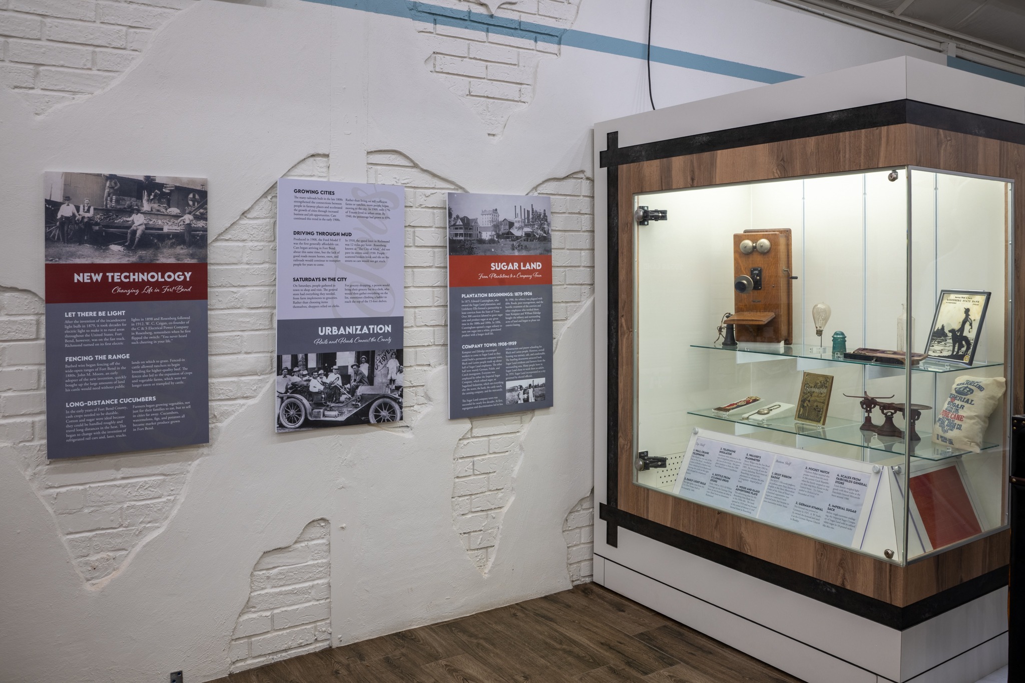A museum exhibit features informational panels on local history and technology, and a glass display case containing vintage objects, photos, and artifacts, all set against a white brick wall.