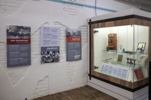Museum exhibit with three informational panels on a white brick wall and a glass display case containing historical artifacts, including photos, books, and vintage objects, on a wooden floor.