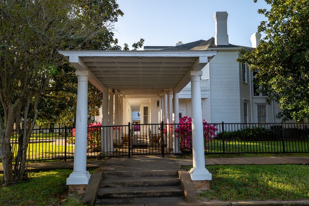 A white wooden house with a covered walkway supported by columns, leading to a black metal gate. Pink flowers and trees surround the building, and sunlight casts shadows on the grass and pavement.