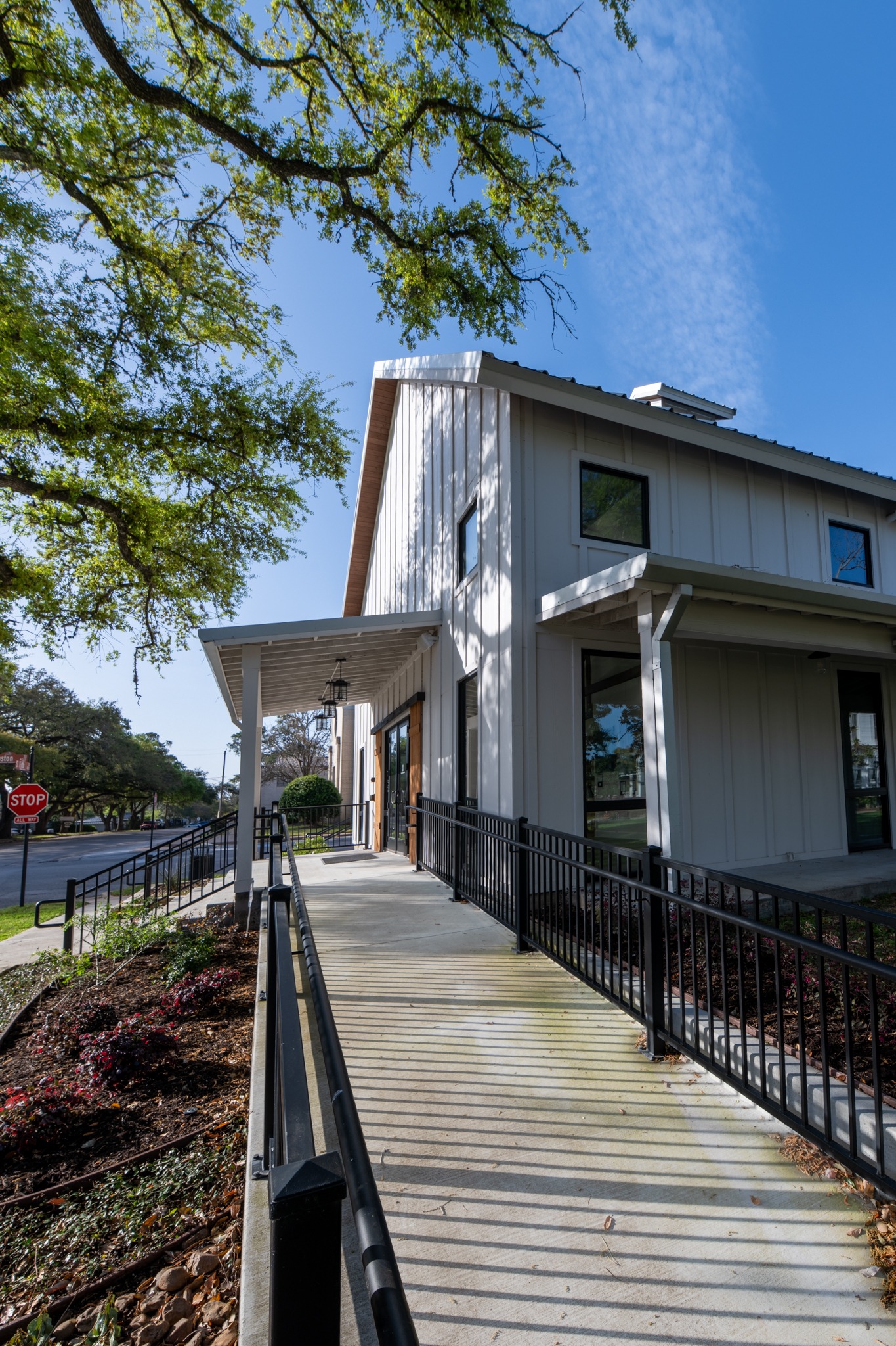 A modern white building with large windows and a sloped roof, featuring an accessible ramp with black railings. Trees with green leaves surround the building, and a stop sign is visible in the background.