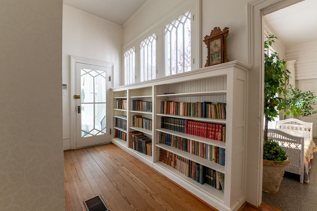 A bright hallway with wooden floors features built-in white bookshelves filled with books, a vintage clock on top, tall windows, and a doorway leading to a cozy room with wicker furniture and a potted plant.