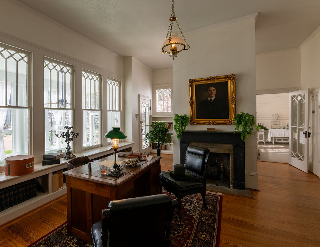 A vintage study with a wooden desk, green desk lamp, two black chairs, a fireplace above which hangs a portrait, large windows with decorative panes, hardwood floors, and houseplants. A sunroom is visible through open doors.