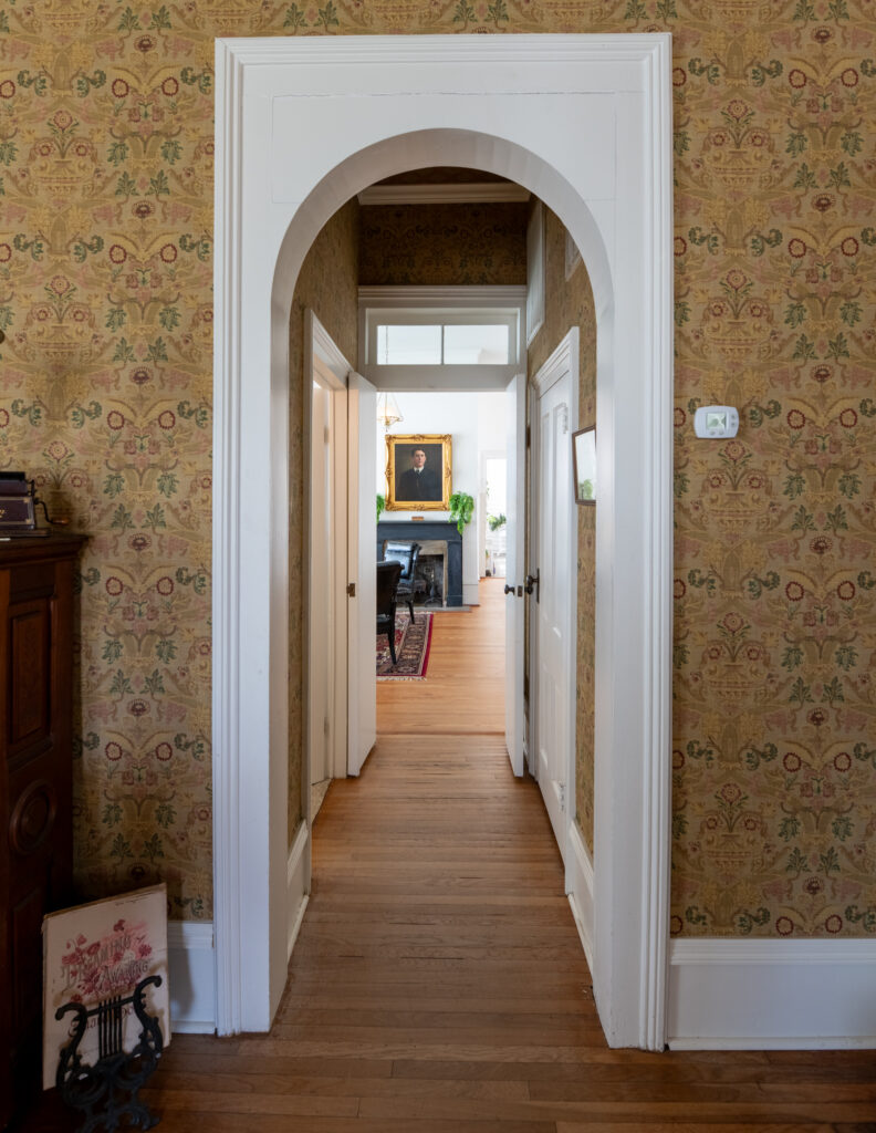 A hallway with wooden floors and patterned wallpaper leads through an arched doorway to a room with a fireplace, plants, and a large framed portrait on the far wall. White trim frames the arch and doors.