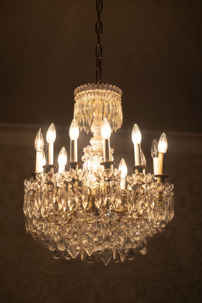 A lit crystal chandelier with multiple candle-shaped bulbs and hanging crystal ornaments, suspended by a chain against a dimly lit background.