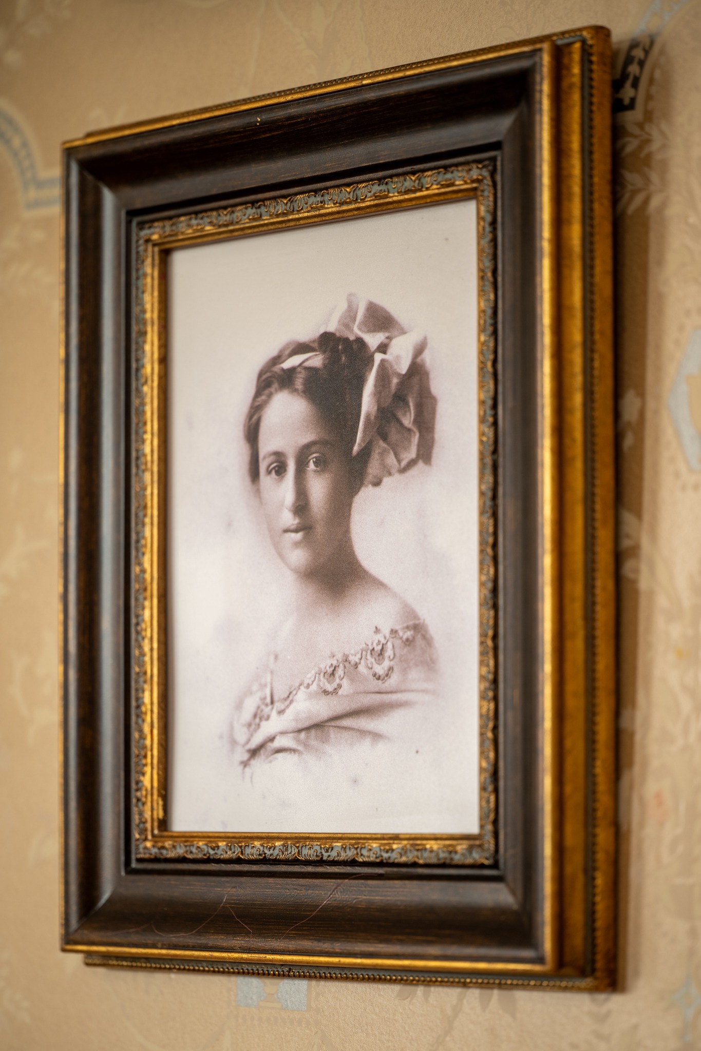A sepia-toned portrait of a woman in an ornate gold and dark wood frame hangs on a patterned wall. The woman has an elaborate bow in her hair and is dressed in an off-the-shoulder, embroidered gown.