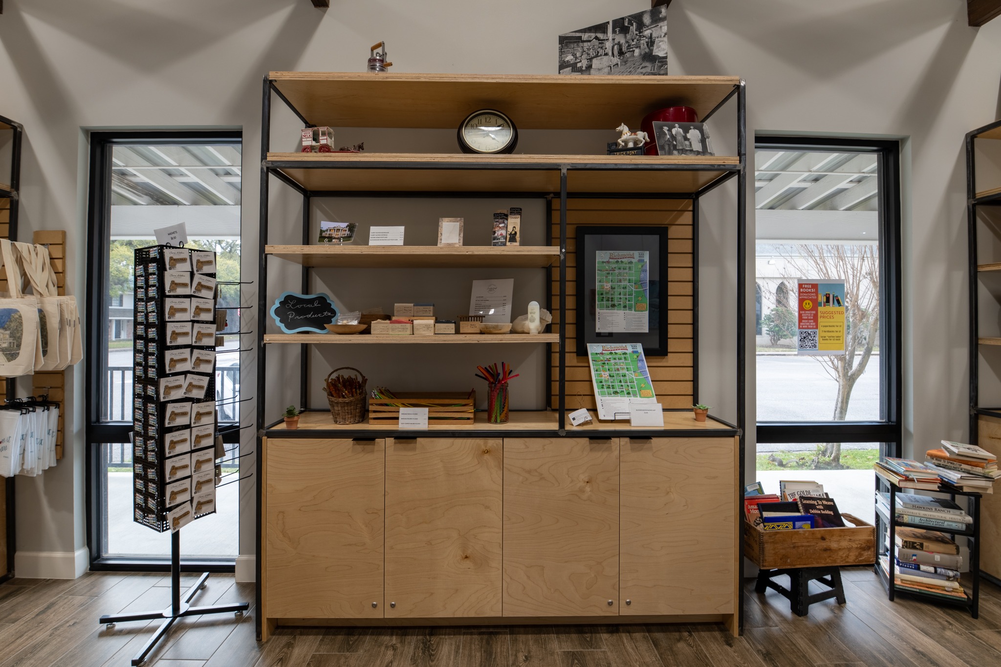 A wooden shelving unit displays various items like books, postcards, small sculptures, framed art, and baskets. Large windows on either side let in daylight, and a rotating postcard rack and a bin of books are nearby.