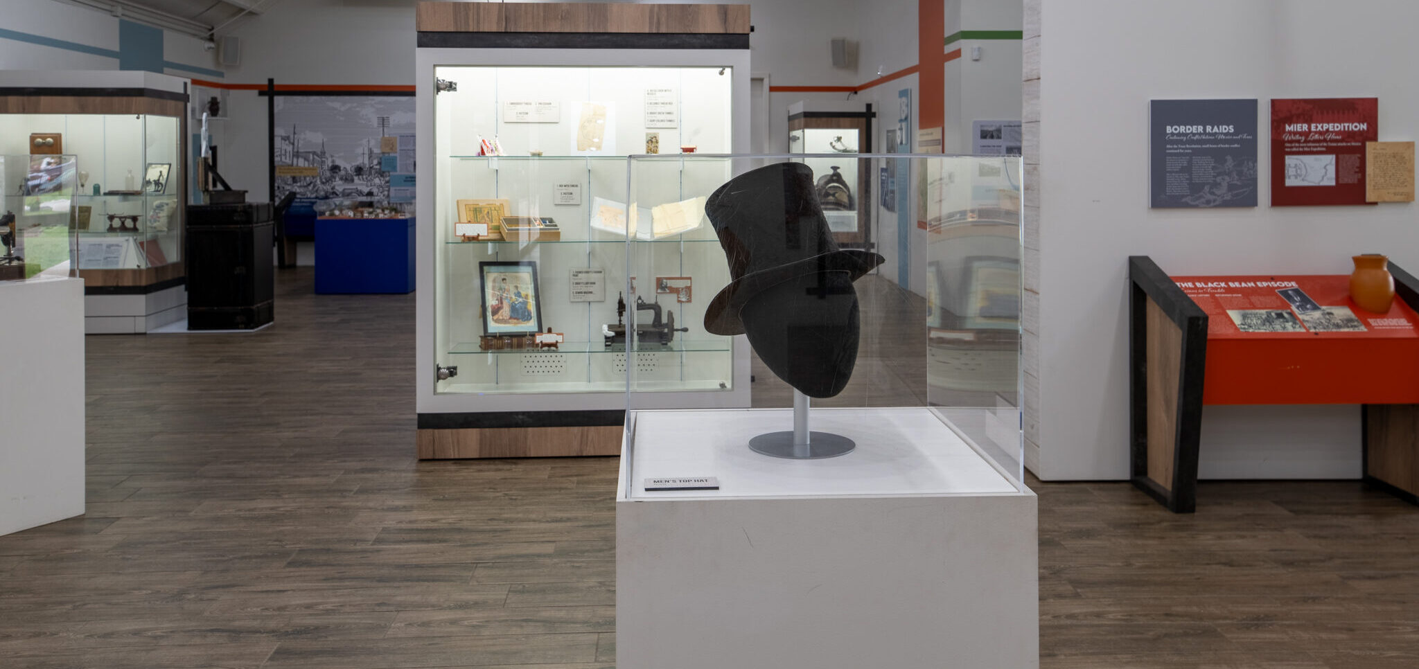 A black top hat is displayed in a glass case at the center of a museum exhibit with wooden floors, informational signs, and display cabinets holding books and artifacts in the background.