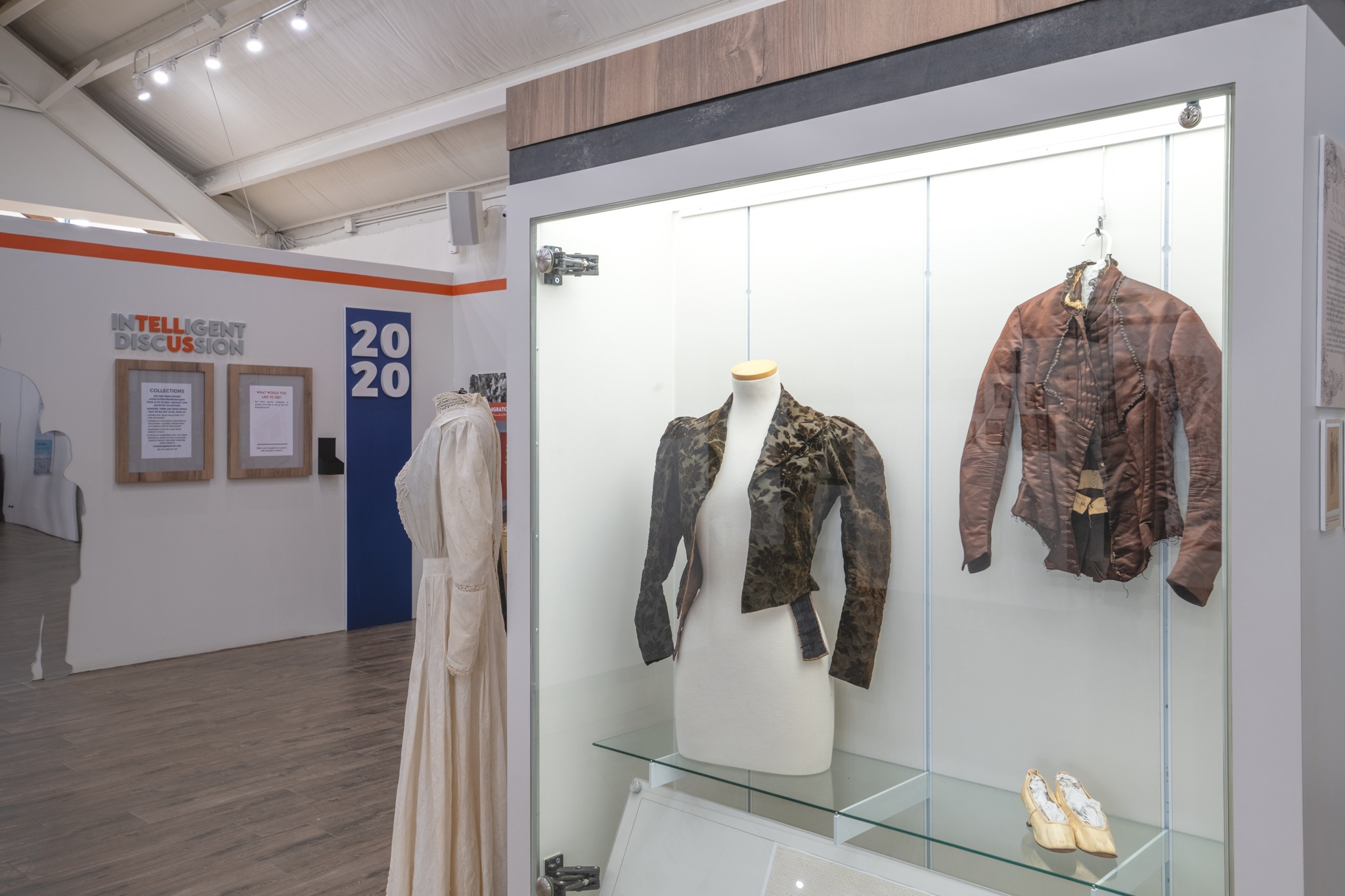 A museum exhibit displays historical clothing, including a cream dress, two brown jackets, and a pair of shoes. In the background, a wall has the text "INTELLIGENT DISCUSSION" and "2020" in bold letters.