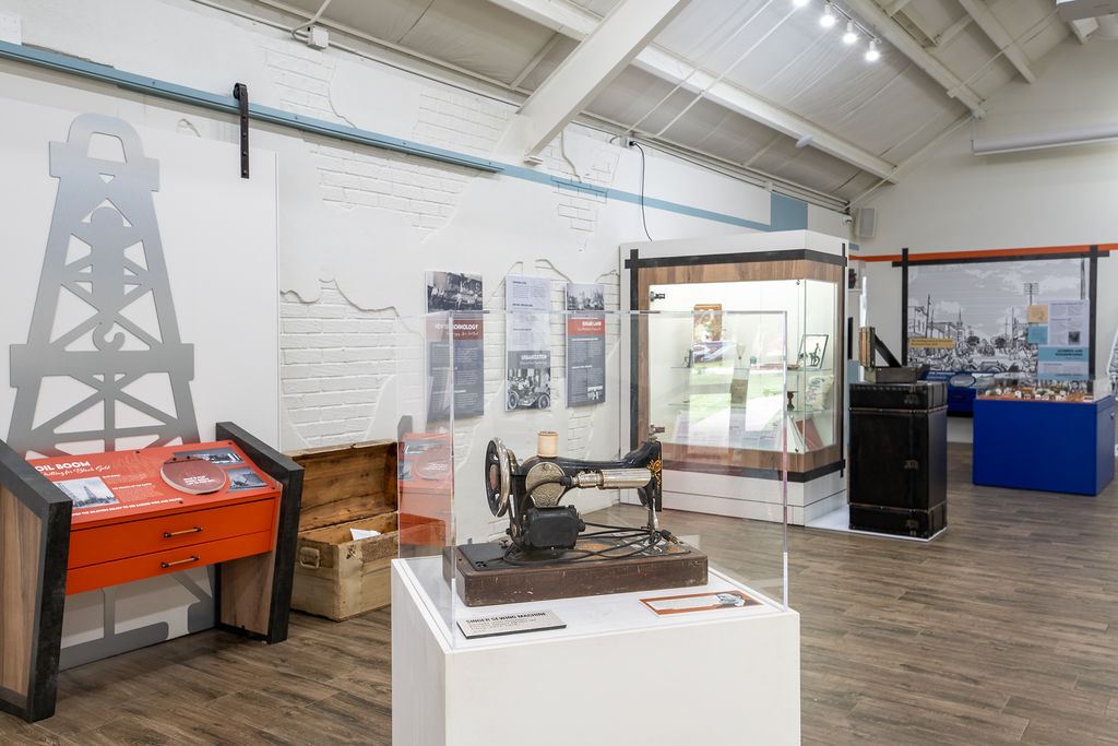 A museum exhibit features a vintage sewing machine in a glass case, an oil derrick display, information panels on the wall, a wooden chest, and various historical artifacts in a well-lit room with wood flooring.