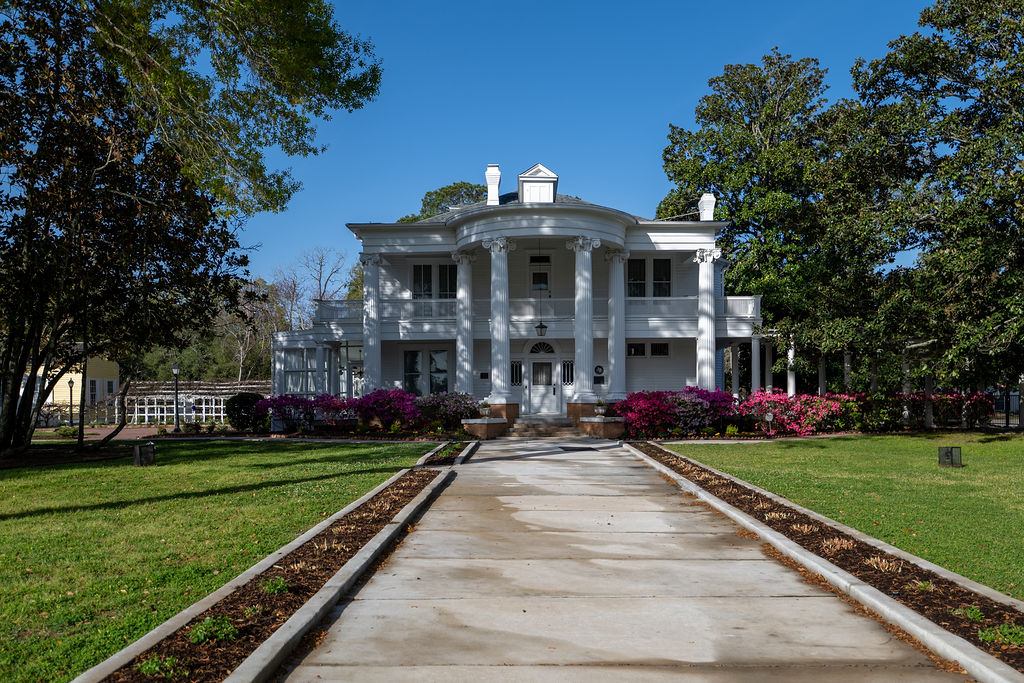 A grand white mansion with tall columns, a large front porch, and blooming pink flowers along the walkway, set among green lawns and tall trees under a clear blue sky.