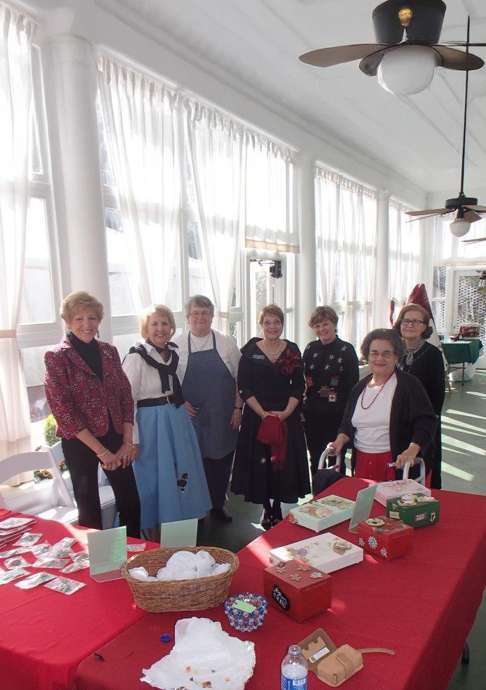 Seven women stand and sit around a table decorated with cards, baskets, and holiday items in a sunlit room with large windows and white curtains. The atmosphere appears cheerful and festive.