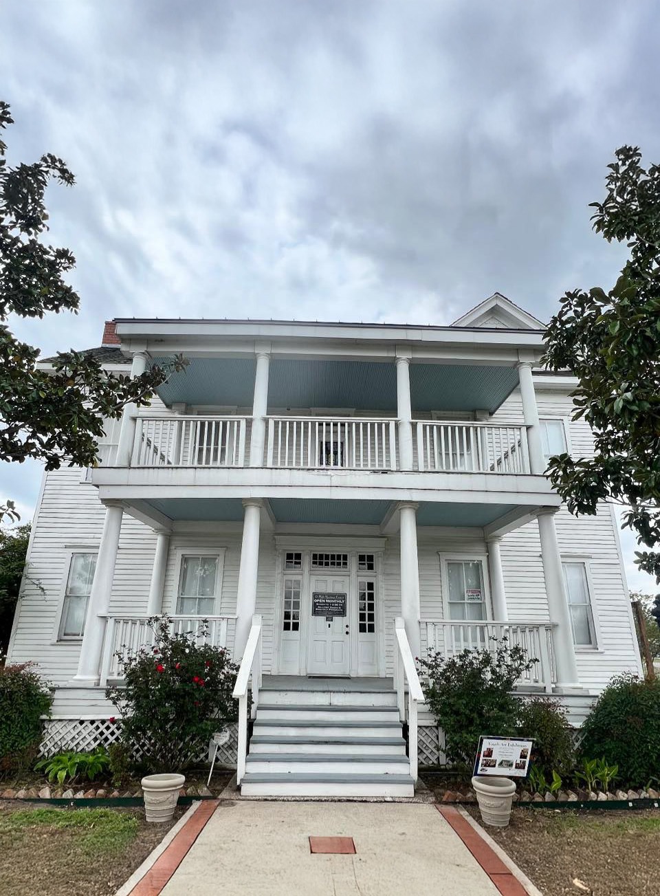 A two-story white house with double front porches, white railings, and stairs leading to the entrance. Bushes and flowers line the walkway, and a cloudy sky is visible above.