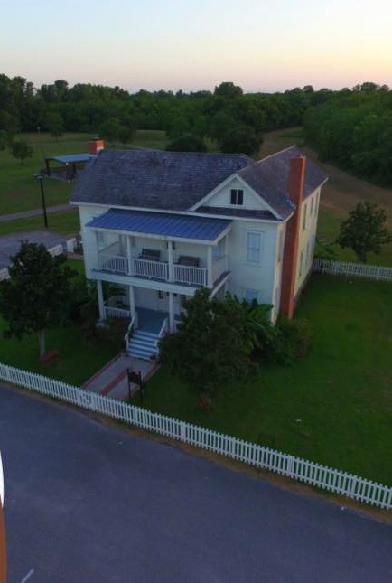 Aerial view of a two-story white house with a wraparound porch, surrounded by a white picket fence and greenery, located at the corner of a paved road and grassy areas.