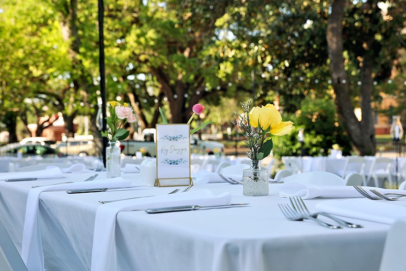 Outdoor table setting with white tablecloths, neatly arranged cutlery, small vases holding colorful flowers, and a decorative menu card, set in a garden with leafy green trees in the background.