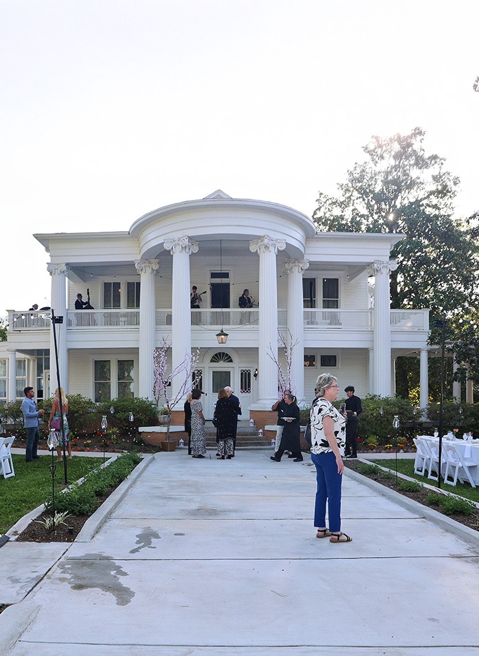 A large white mansion with tall columns, a balcony, and several people gathered outside on the driveway and balconies during a daytime event. There are tables set up with white tablecloths on the lawn.