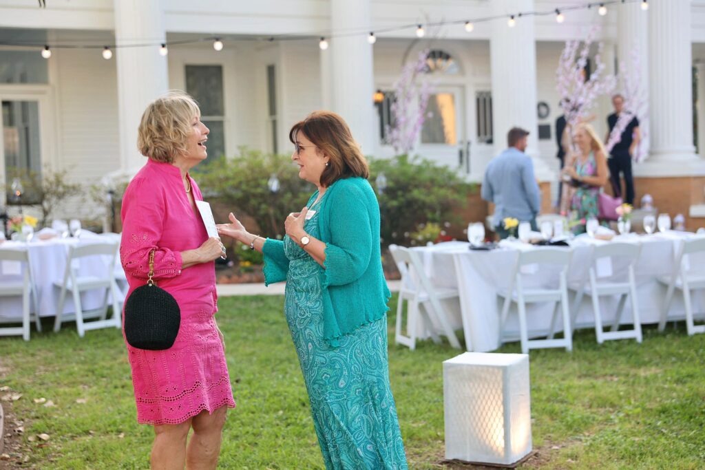 Two women stand and chat on a lawn at an outdoor event with white-clothed tables and string lights, in front of a large white building with columns. Other guests are visible in the background.