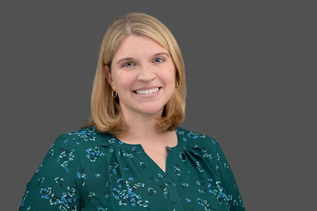 Woman with straight blonde hair, wearing a green blouse with a blue floral pattern, smiles at the camera against a plain gray background.
