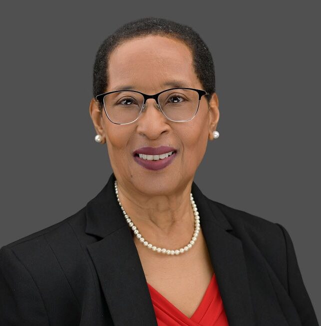 Portrait of a smiling woman wearing glasses, pearl earrings, a pearl necklace, a red blouse, and a black blazer, against a plain gray background.