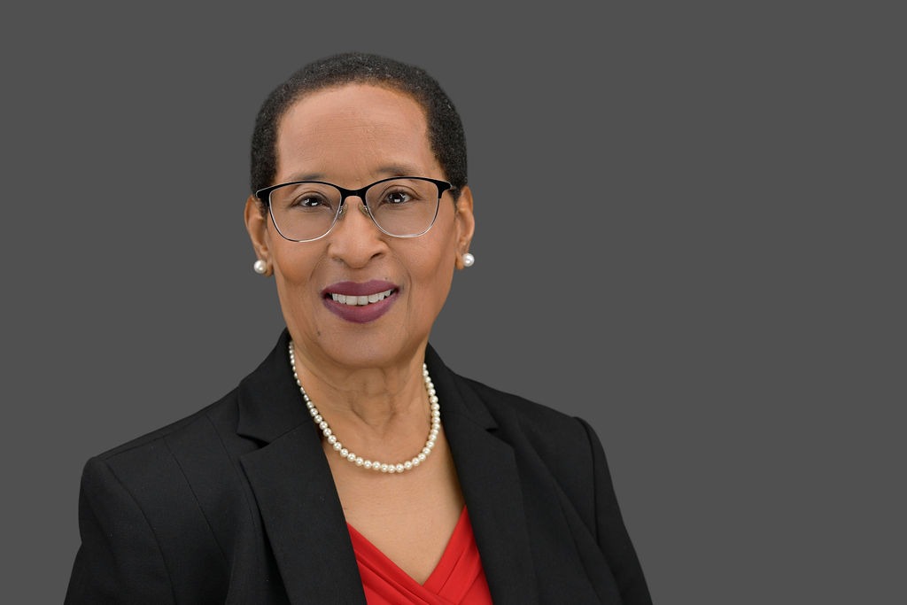 Portrait of a smiling woman wearing glasses, pearl earrings, a pearl necklace, a red blouse, and a black blazer, against a plain gray background.