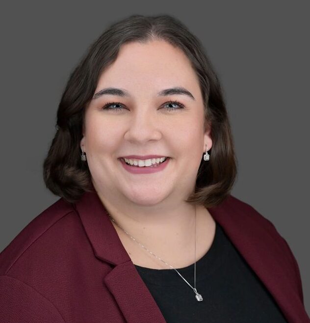 A woman with short brown hair smiles, wearing a burgundy blazer, black top, and a silver necklace with a pendant, against a plain gray background.