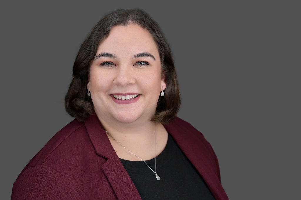 A woman with short brown hair smiles, wearing a burgundy blazer, black top, and a silver necklace with a pendant, against a plain gray background.