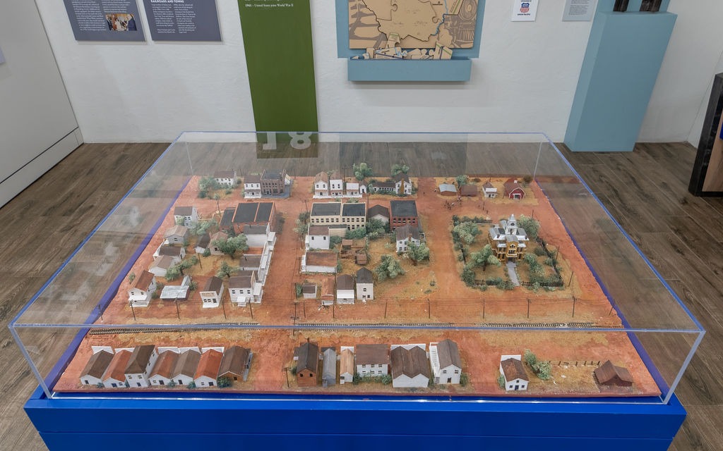 A museum display features a detailed scale model of a small town with miniature buildings, streets, and trees, all enclosed in a clear glass case set on a blue pedestal.