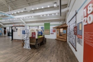 A museum exhibit features a covered wagon with a life-size cutout of a person beside it, informative panels on the walls, and a small log cabin display in a well-lit room with wood flooring.