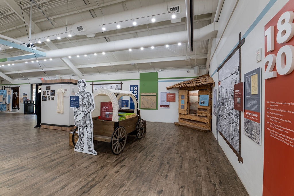 A museum exhibit features a covered wagon with a life-size cutout of a person beside it, informative panels on the walls, and a small log cabin display in a well-lit room with wood flooring.