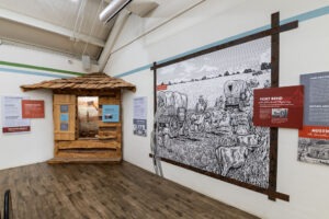 Museum exhibit featuring a wooden frontier cabin facade and a large black-and-white mural of covered wagons crossing a field, with informational panels about Texas history on the surrounding walls.